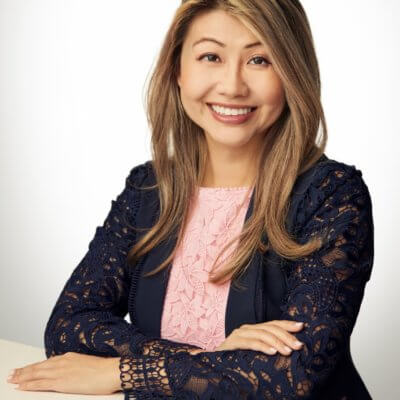 Joanna Hu, an Asian woman with light brown hair, wears a navy blazer over a light pink shirt with her hand resting on a white shelf and smiling.