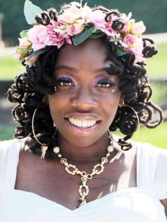 A headshot of Tanesha, a Black woman with curly hair, smiling in a white top with a pink and white flower crown, and wearing jewelry.